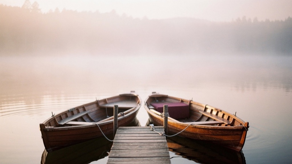 Two wooden rowing boats side by side at a narrow jetty on a still lake, one holding a dark red cushion, a wooded shore behind them in morning mist, soft pinkish light